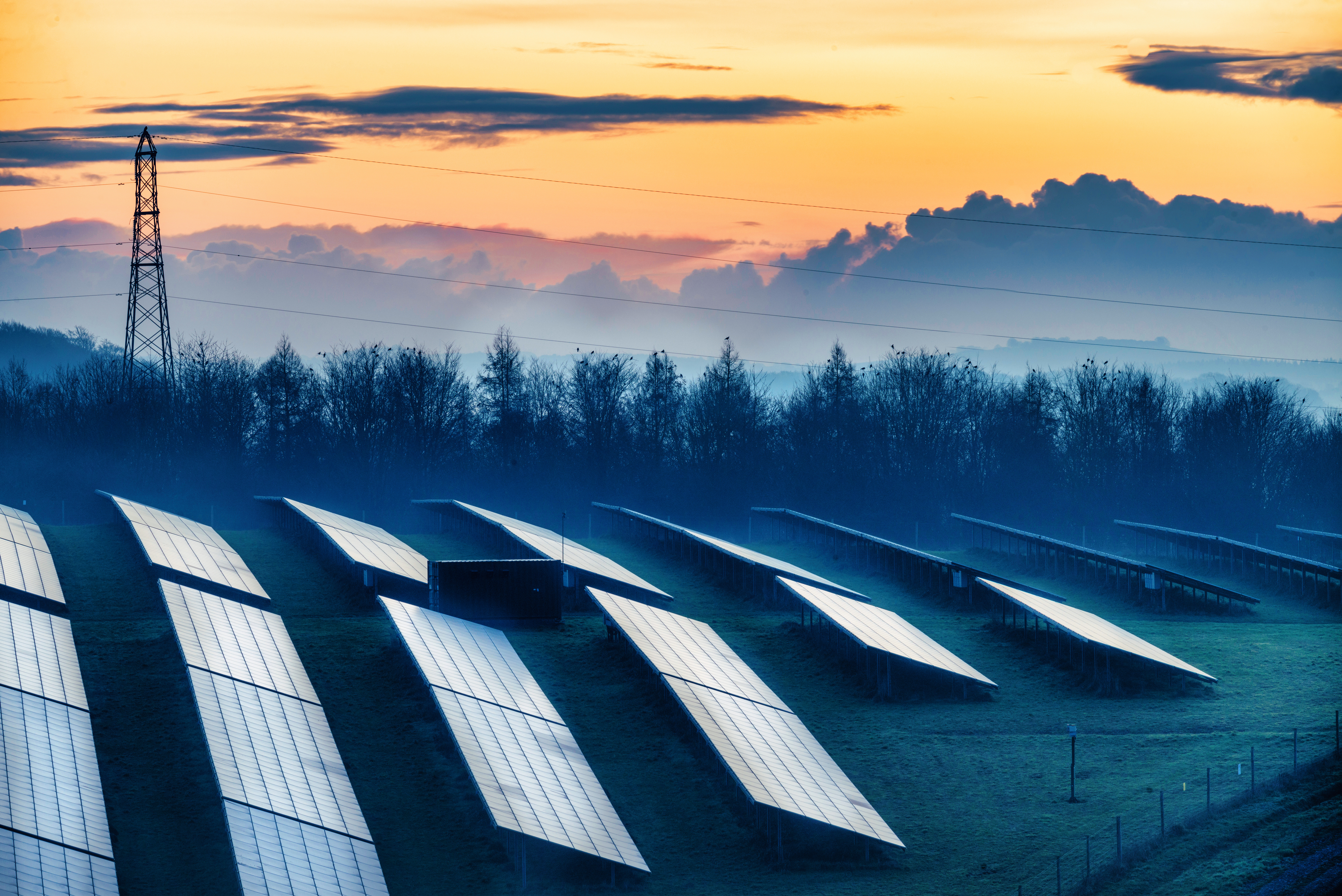 Multiple solar panels reflecting the fading winter sunlight at dusk, and glowing afterlight in the sky beyond, in the rural countryside.